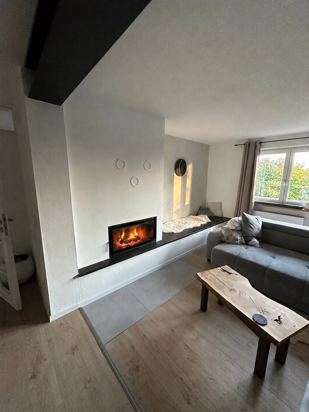 Breite weiße Ofenwand mit länglicher Feuerraumscheibe und Bank neben dem Sofa. Feuer mit KI bearbeitet.