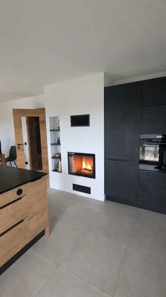 Deckenhoher Kamin mit breiter Scheibe, schwarzen Gittern und Regal in Velden. Feuer mit KI bearbeitet.